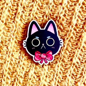 May include: Black cat with pink ears and a pink bow tie. The cat has a white face with black eyes and a black nose.