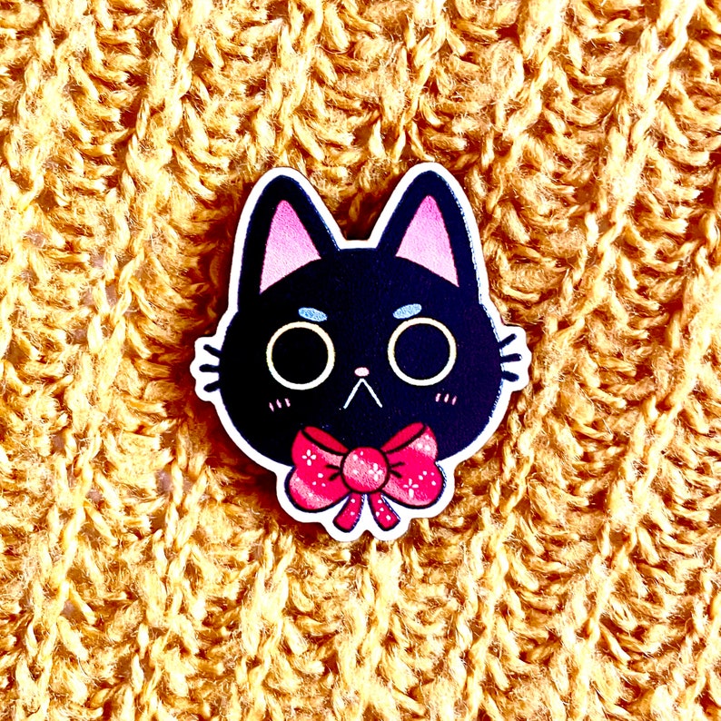 May include: Black cat with pink ears and a pink bow tie. The cat has a white face with black eyes and a black nose.