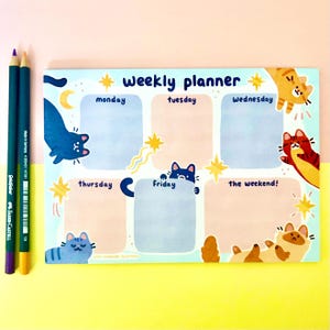 May include: A light blue weekly planner with cat illustrations and the text "weekly planner". The planner has sections for each day of the week, with cat graphics and star accents. Two colored pencils are on the left.
