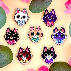 Op de afbeelding: Zeven kleurrijke kattengezichtspinnen met verschillende gekleurde vacht en strikjes. De pinnen bevinden zich op een lichtgele achtergrond met bloemen en bladeren.
