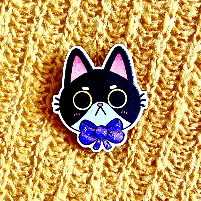 May include: A black and white cat with pink ears and a purple bow tie. The cat is wearing a white collar and has a cute expression on its face.