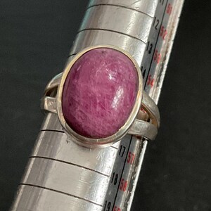 Pu&ograve; includere: Un anello in argento con una grande pietra preziosa cabochon di forma ovale color rosa intenso. L'anello &egrave; posizionato su un mandrino in metallo, con la fascia che poggia tra i segni 14 e 15 dello strumento di misurazione, indicandone la misura.