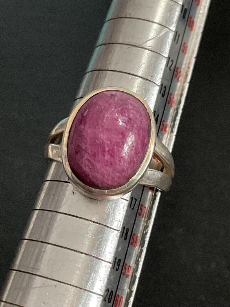 Pu&ograve; includere: Un anello in argento con una grande pietra preziosa cabochon di forma ovale color rosa intenso. L'anello &egrave; posizionato su un mandrino in metallo, con la fascia che poggia tra i segni 14 e 15 dello strumento di misurazione, indicandone la misura.