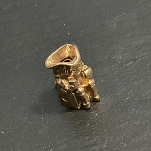 K&ouml;nnte beinhalten: Ein kleiner, dreidimensionaler goldfarbener Anh&auml;nger in Form einer Figur mit breitkrempigem Hut. Der Anh&auml;nger verf&uuml;gt &uuml;ber eine hohle &Ouml;ffnung an der Oberseite und detaillierte, geformte Merkmale einer bekleideten Person mit metallischem Glanz.