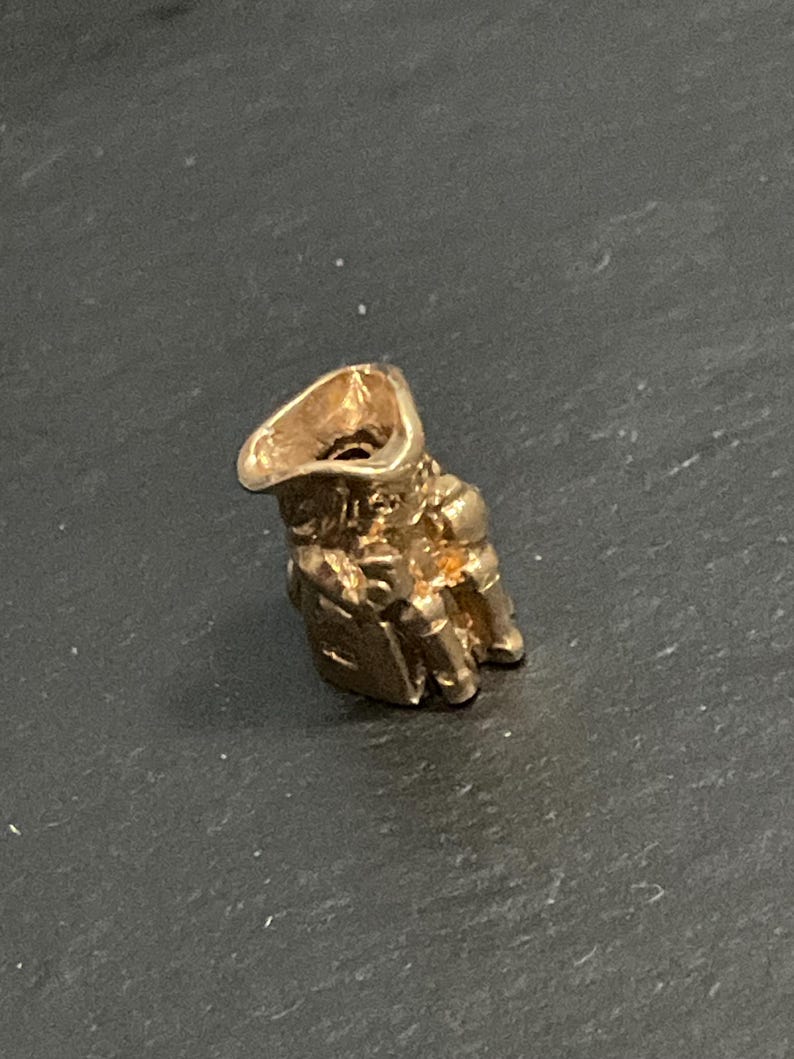 K&ouml;nnte beinhalten: Ein kleiner, dreidimensionaler goldfarbener Anh&auml;nger in Form einer Figur mit breitkrempigem Hut. Der Anh&auml;nger verf&uuml;gt &uuml;ber eine hohle &Ouml;ffnung an der Oberseite und detaillierte, geformte Merkmale einer bekleideten Person mit metallischem Glanz.