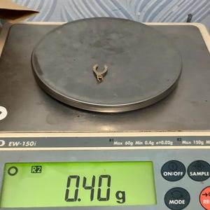 K&ouml;nnte beinhalten: Ein kleiner, metallischer Anh&auml;nger in Hufeisenform liegt auf der runden Plattform einer digitalen Waage. Das gr&uuml;n beleuchtete Display der Waage zeigt deutlich ein Gewicht von 0,40 Gramm f&uuml;r das Objekt an, das eine kleine &Ouml;se besitzt.