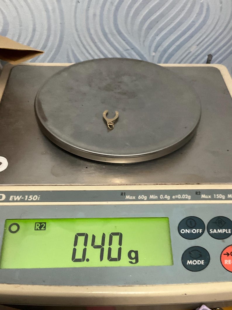 K&ouml;nnte beinhalten: Ein kleiner, metallischer Anh&auml;nger in Hufeisenform liegt auf der runden Plattform einer digitalen Waage. Das gr&uuml;n beleuchtete Display der Waage zeigt deutlich ein Gewicht von 0,40 Gramm f&uuml;r das Objekt an, das eine kleine &Ouml;se besitzt.