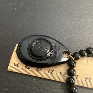 May include: A black vintage necklace with a teardrop-shaped pendant and a beaded strand. The pendant features a floral design and is approximately 2 inches long. The necklace is displayed with a wooden ruler.