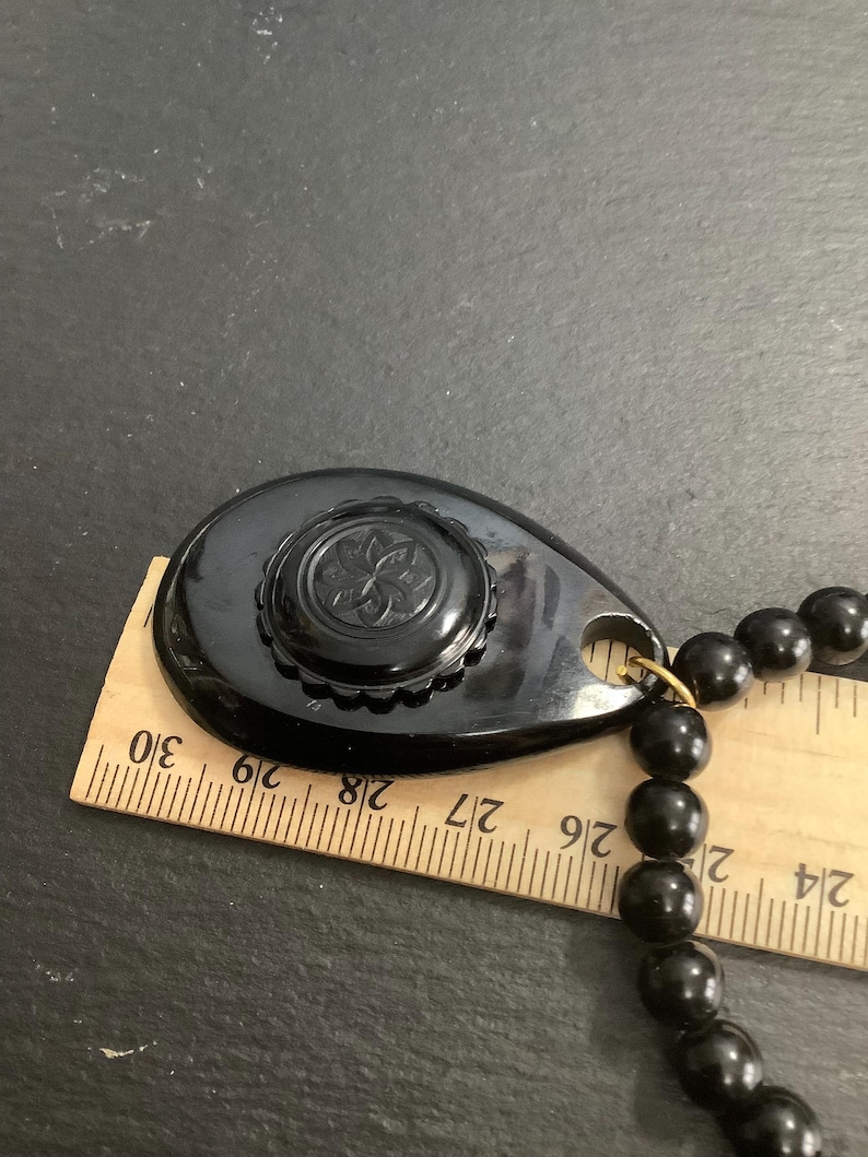 May include: A black vintage necklace with a teardrop-shaped pendant and a beaded strand. The pendant features a floral design and is approximately 2 inches long. The necklace is displayed with a wooden ruler.