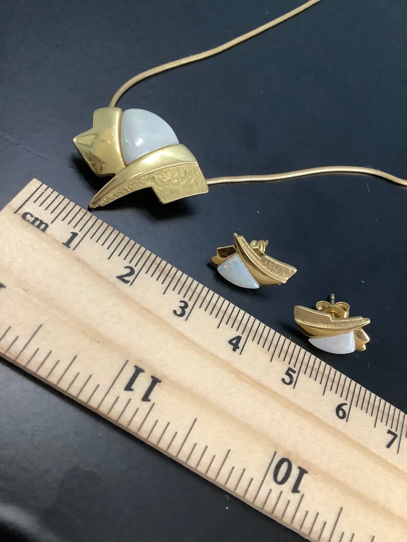 Op de afbeelding: Een gouden ketting en oorbellen set met een witte opaal steen. De ketting heeft een geometrisch ontwerp en de oorbellen zijn eenvoudige studs.