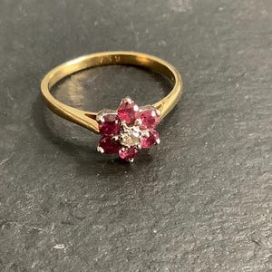 May include: A gold ring with a floral design, set with red gemstones and a central diamond. The ring's band is slender, and the setting is on a dark surface. The ring is marked with the number 750.