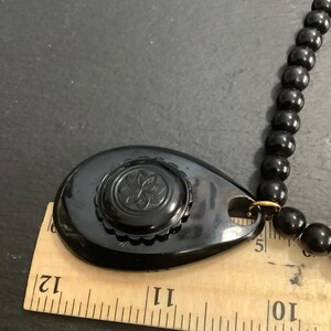 May include: A vintage black necklace featuring a large, teardrop-shaped pendant with a floral design. The pendant is connected to a beaded necklace. The ruler shows the pendant is approximately 2.5 inches long.