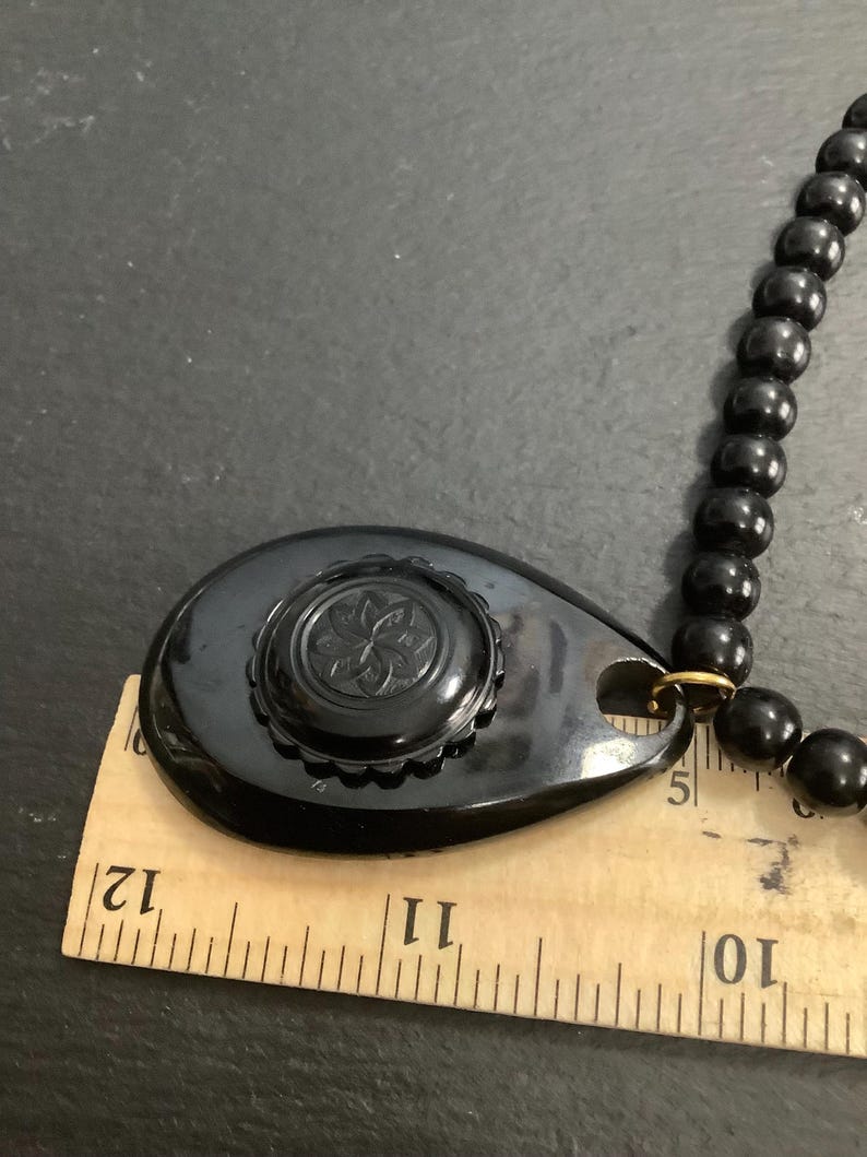 May include: A vintage black necklace featuring a large, teardrop-shaped pendant with a floral design. The pendant is connected to a beaded necklace. The ruler shows the pendant is approximately 2.5 inches long.