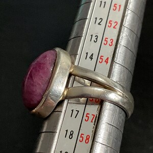 Pu&ograve; includere: Un anello in argento con una pietra liscia di forma ovale color magenta intenso incastonata in una montatura. L'anello &egrave; posizionato su un mandrino in metallo, con la fascia che poggia tra le taglie 15 e 16, indicando una misura di circa 15,5.