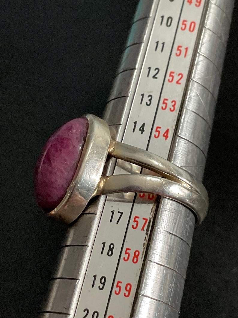Pu&ograve; includere: Un anello in argento con una pietra liscia di forma ovale color magenta intenso incastonata in una montatura. L'anello &egrave; posizionato su un mandrino in metallo, con la fascia che poggia tra le taglie 15 e 16, indicando una misura di circa 15,5.