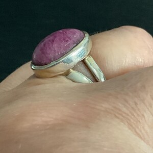 Pu&ograve; includere: Un anello in argento con una grande pietra preziosa color magenta di forma ovale, taglio cabochon, incastonata in una lunetta di metallo liscio. Il gambo dell'anello &egrave; diviso in due segmenti d'argento distinti collegati alla base dell'incastonatura.