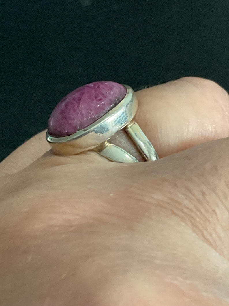 Pu&ograve; includere: Un anello in argento con una grande pietra preziosa color magenta di forma ovale, taglio cabochon, incastonata in una lunetta di metallo liscio. Il gambo dell'anello &egrave; diviso in due segmenti d'argento distinti collegati alla base dell'incastonatura.