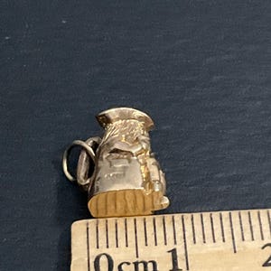 K&ouml;nnte beinhalten: Ein goldfarbener Metallanh&auml;nger in Form eines kleinen Kruges oder einer Kanne mit Henkel und Ausguss. Der Anh&auml;nger liegt neben einem Holzlineal, das zeigt, dass das Objekt eine Gesamth&ouml;he von etwa 1,5 Zentimetern aufweist.