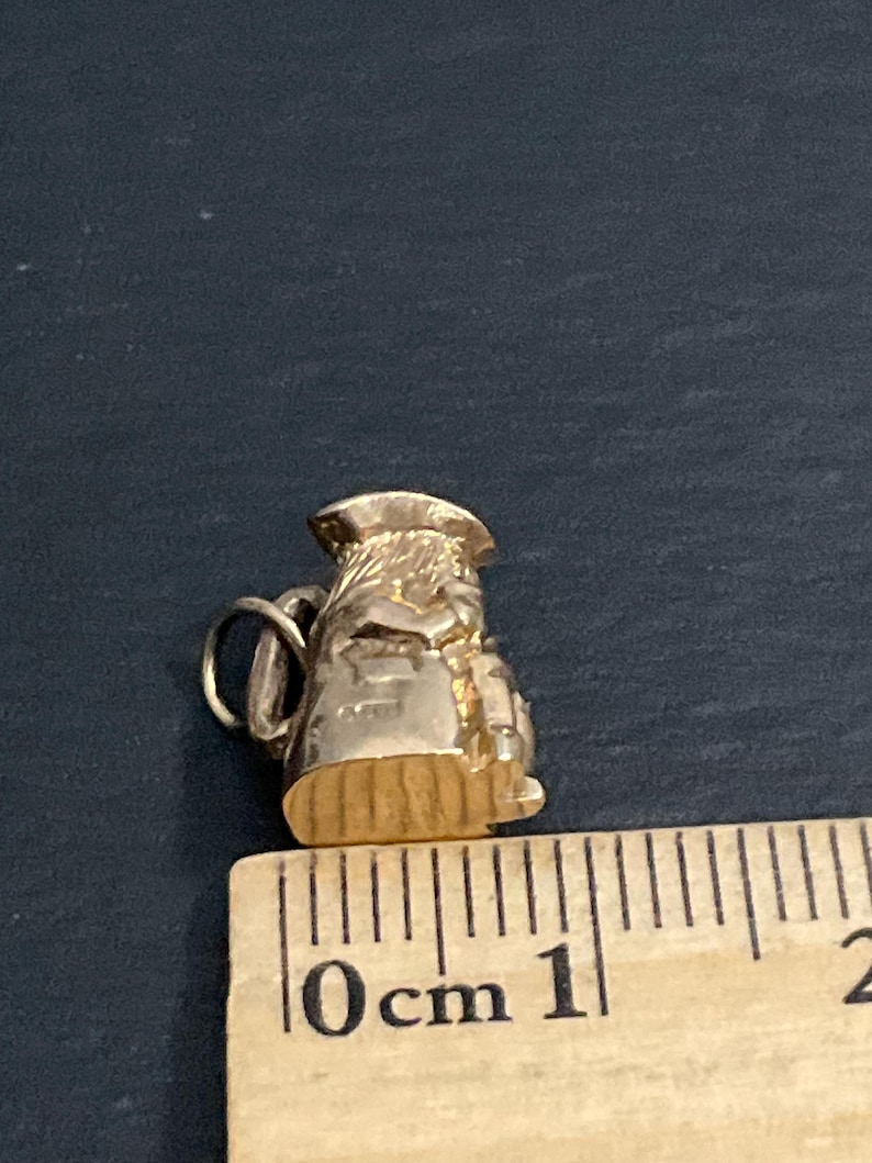 K&ouml;nnte beinhalten: Ein goldfarbener Metallanh&auml;nger in Form eines kleinen Kruges oder einer Kanne mit Henkel und Ausguss. Der Anh&auml;nger liegt neben einem Holzlineal, das zeigt, dass das Objekt eine Gesamth&ouml;he von etwa 1,5 Zentimetern aufweist.