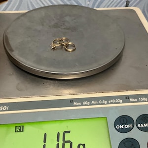 K&ouml;nnte beinhalten: Zwei goldfarbene Ringe, einer davon mit einem kleinen klaren Stein besetzt, liegen auf einer digitalen Waage, die ein Gewicht von 1,16 Gramm anzeigt. Die Waage hat eine runde Wiegefl&auml;che und ein gr&uuml;nes Digitaldisplay f&uuml;r den Messwert.