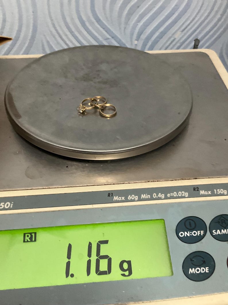 K&ouml;nnte beinhalten: Zwei goldfarbene Ringe, einer davon mit einem kleinen klaren Stein besetzt, liegen auf einer digitalen Waage, die ein Gewicht von 1,16 Gramm anzeigt. Die Waage hat eine runde Wiegefl&auml;che und ein gr&uuml;nes Digitaldisplay f&uuml;r den Messwert.