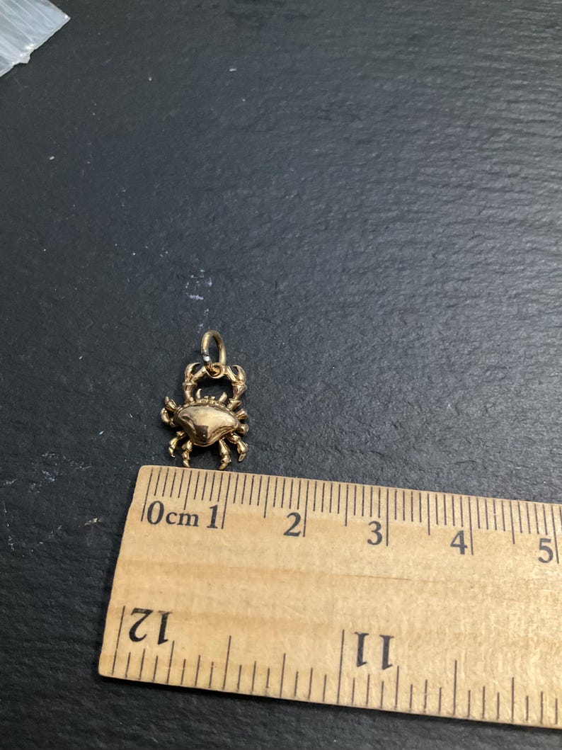 K&ouml;nnte beinhalten: Ein goldfarbener Krabbenanh&auml;nger mit einem kleinen Verbindungsring an der Oberseite. Der Anh&auml;nger liegt &uuml;ber einem Holzlineal, das zeigt, dass die Krabbe selbst eine Breite und H&ouml;he von etwa 2 cm aufweist.