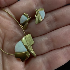 Op de afbeelding: Een gouden ketting en oorbellen set met witte opaalstenen. De ketting heeft een hanger met een geometrisch ontwerp en de oorbellen zijn eenvoudige studs.