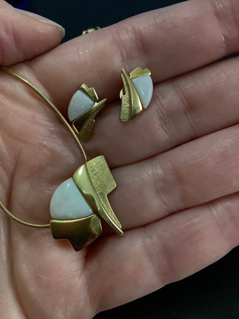 Op de afbeelding: Een gouden ketting en oorbellen set met witte opaalstenen. De ketting heeft een hanger met een geometrisch ontwerp en de oorbellen zijn eenvoudige studs.