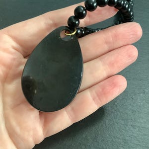 May include: A black teardrop-shaped pendant necklace with a beaded chain. The pendant is smooth and glossy, with a small gold-colored ring at the top. The necklace is made of black beads.