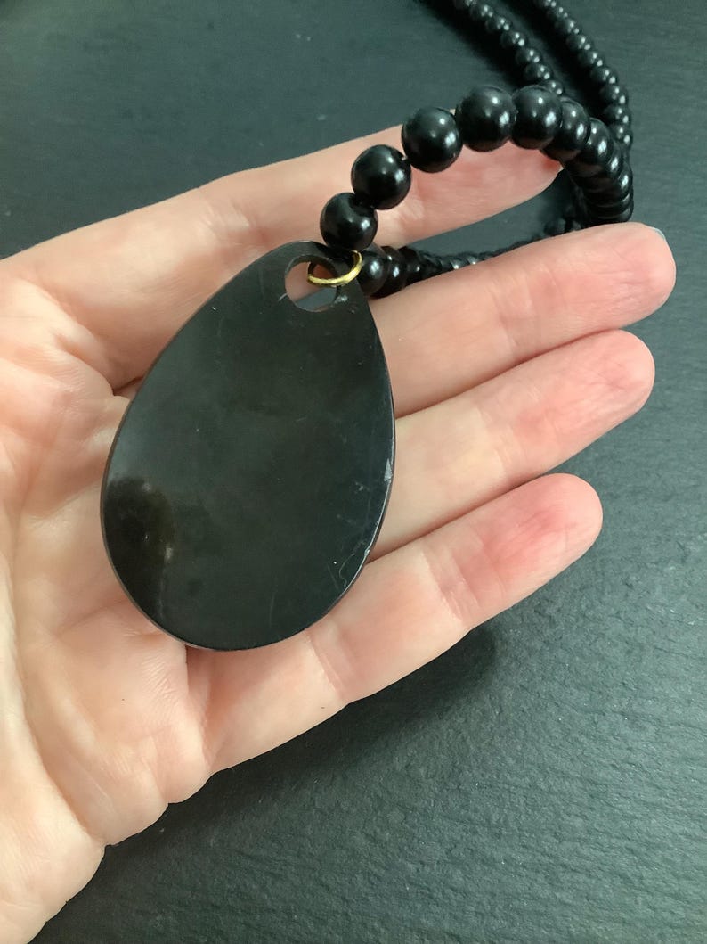 May include: A black teardrop-shaped pendant necklace with a beaded chain. The pendant is smooth and glossy, with a small gold-colored ring at the top. The necklace is made of black beads.