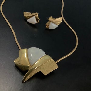 Op de afbeelding: Een gouden ketting met een hanger met een witte opaal-steen in een geometrisch ontwerp. De ketting wordt geleverd met bijpassende oorbellen met witte opaal-stenen.