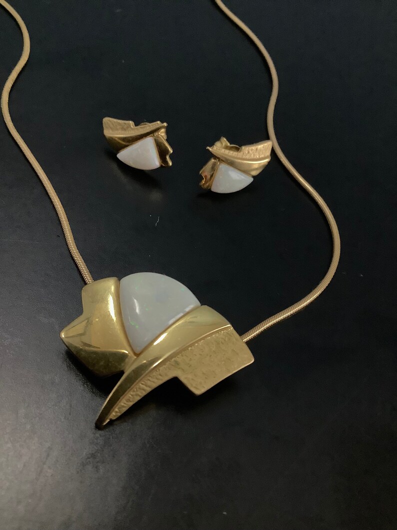 Op de afbeelding: Een gouden ketting met een hanger met een witte opaal-steen in een geometrisch ontwerp. De ketting wordt geleverd met bijpassende oorbellen met witte opaal-stenen.