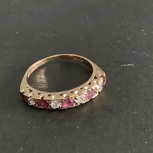 Könnte beinhalten: Ein goldener Ring mit abwechselnd rosafarbenen und klaren Edelsteinen. Der Ring hat ein zartes Band, und die Edelsteine sind gleichmäßig verteilt. Der Ring ist vor einem dunklen Hintergrund platziert.