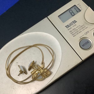 Op de afbeelding: Een gouden ketting met een hanger met een witte steen en twee kleinere witte stenen aan de zijkanten. De ketting ligt op een digitale weegschaal die 8,1 gram aangeeft.