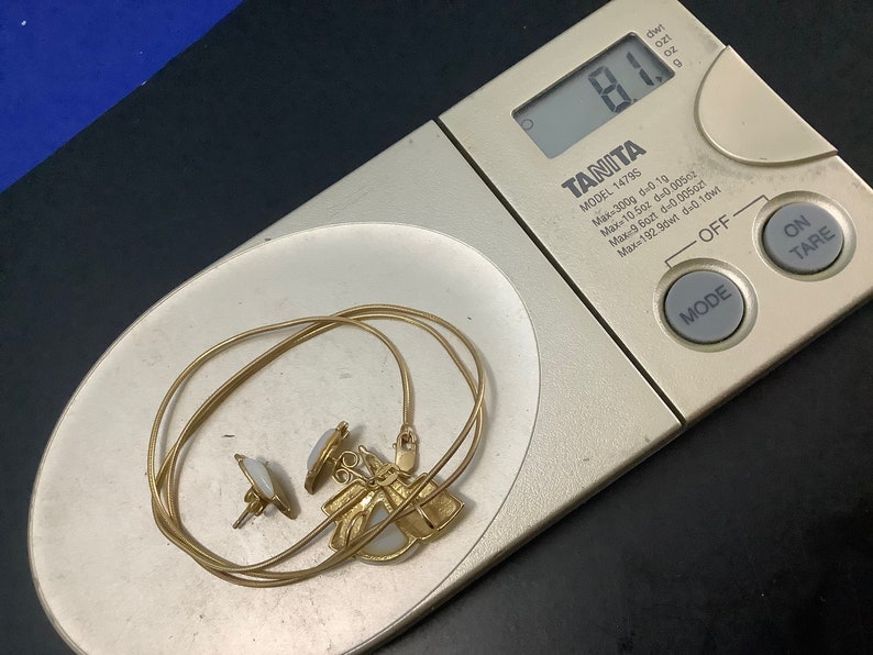 Op de afbeelding: Een gouden ketting met een hanger met een witte steen en twee kleinere witte stenen aan de zijkanten. De ketting ligt op een digitale weegschaal die 8,1 gram aangeeft.