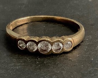 Puede incluir: Un anillo de oro con cinco piedras preciosas transparentes. El anillo tiene un diseño clásico con un aspecto ligeramente desgastado. Las piedras preciosas, probablemente diamantes, son redondas. El anillo está sobre un fondo oscuro.