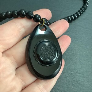 May include: A black beaded necklace with a teardrop-shaped pendant. The pendant features a carved floral design within a circular frame. The necklace is a dark, glossy black.