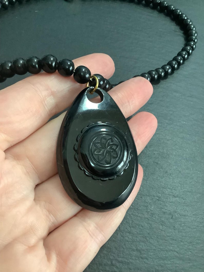 May include: A black beaded necklace with a teardrop-shaped pendant. The pendant features a carved floral design within a circular frame. The necklace is a dark, glossy black.