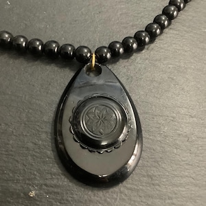 May include: A black beaded necklace with a teardrop pendant. The pendant features a floral design in the center and is attached to the necklace with a gold-colored loop. The beads are small and round.
