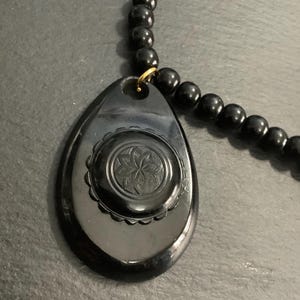 May include: A black beaded necklace with a teardrop-shaped pendant. The pendant features a floral design in the center and is attached to the necklace with a gold-colored ring. The beads are round and evenly spaced.