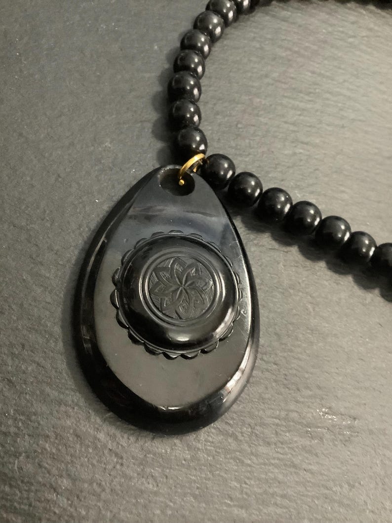 May include: A black beaded necklace with a teardrop-shaped pendant. The pendant features a floral design in the center and is attached to the necklace with a gold-colored ring. The beads are round and evenly spaced.