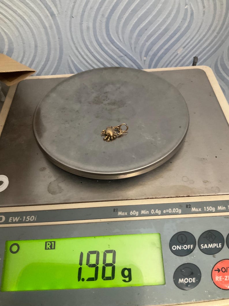 K&ouml;nnte beinhalten: Ein kleiner goldfarbener Krabbenanh&auml;nger mit einem runden Verbindungsring, der auf einer digitalen Waage liegt. Das Display der Waage zeigt ein Gewicht von 1,98 Gramm an. Das Ger&auml;t ist mit EW-150i beschriftet und hat eine Kapazit&auml;t von 150 Gramm.
