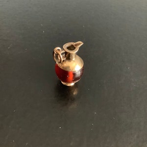 May include: A gold-coloured charm in the shape of a small pitcher with a red glass-like body. The charm has a loop for attaching to a necklace or bracelet.