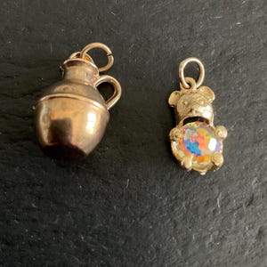 Könnte beinhalten: Zwei Anhänger aus goldfarbenem Metall. Ein Anhänger ist ein kleiner Krug mit Henkel und einem runden Verbindungsring an der Oberseite. Der zweite Anhänger ist eine Bärenfigur mit einem runden, schillernden, mehrfarbigen facettierten Stein.