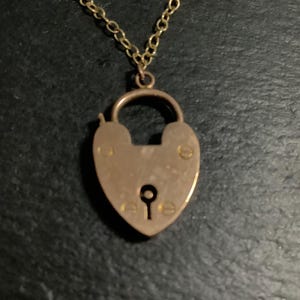 Könnte beinhalten: Eine goldfarbene herzförmige Vorhängeschloss-Anhänger-Halskette. Der Anhänger hat ein Schlüsselloch und ist an einer filigranen Kette befestigt. Die Halskette wird auf einer dunklen Oberfläche präsentiert.