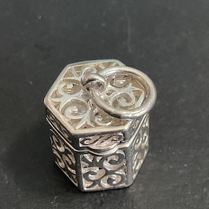 May include: A hexagonal silver charm featuring an intricate openwork filigree pattern on the lid and sides. The charm has a polished metallic finish and includes a circular jump ring attached to the top, allowing it to be suspended from a chain.