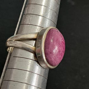 Pu&ograve; includere: Un anello in argento con una pietra preziosa rosa scuro, liscia e di forma ovale, incastonata in una montatura. L'anello &egrave; posizionato su un mandrino in metallo con linee orizzontali, mostrando un design a fascia divisa con finitura lucida.