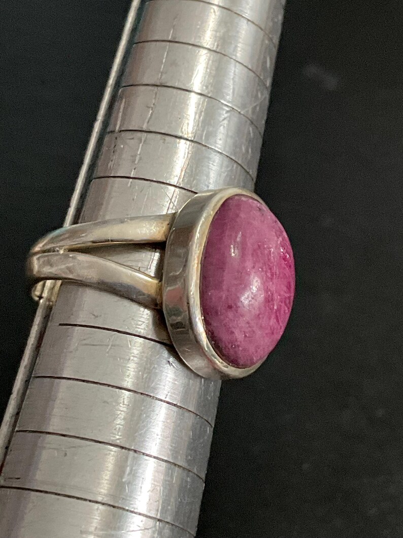 Pu&ograve; includere: Un anello in argento con una pietra preziosa rosa scuro, liscia e di forma ovale, incastonata in una montatura. L'anello &egrave; posizionato su un mandrino in metallo con linee orizzontali, mostrando un design a fascia divisa con finitura lucida.