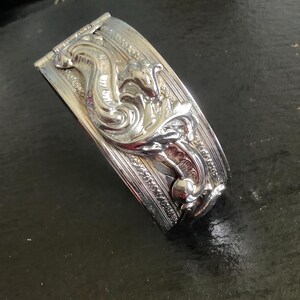 May include: Silver-toned metal cuff bracelet with an ornate design. The bracelet features a detailed relief of a mythical creature, possibly a dragon or serpent, amidst swirling patterns. The cuff is rectangular with a hinged closure.