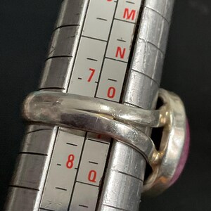 Pu&ograve; includere: Un anello in argento con una pietra rosa posizionato su un mandrino per anelli in metallo. La fascia dell'anello si allinea con la scala di misurazione, che presenta segni numerici e alfabetici per una misurazione precisa della misura.